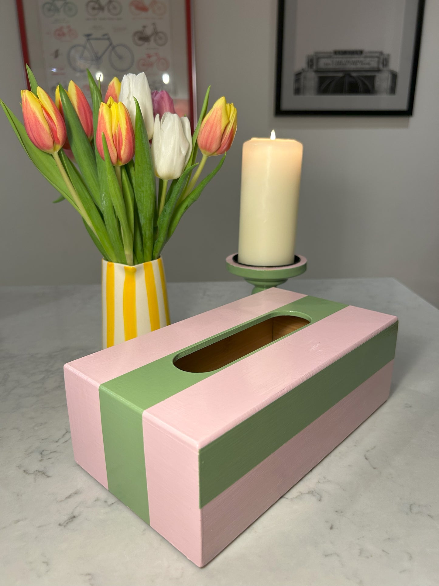 A hand painted pink and green striped tissue box is displayed on a table, accompanied by a vase with tulips and a candle.