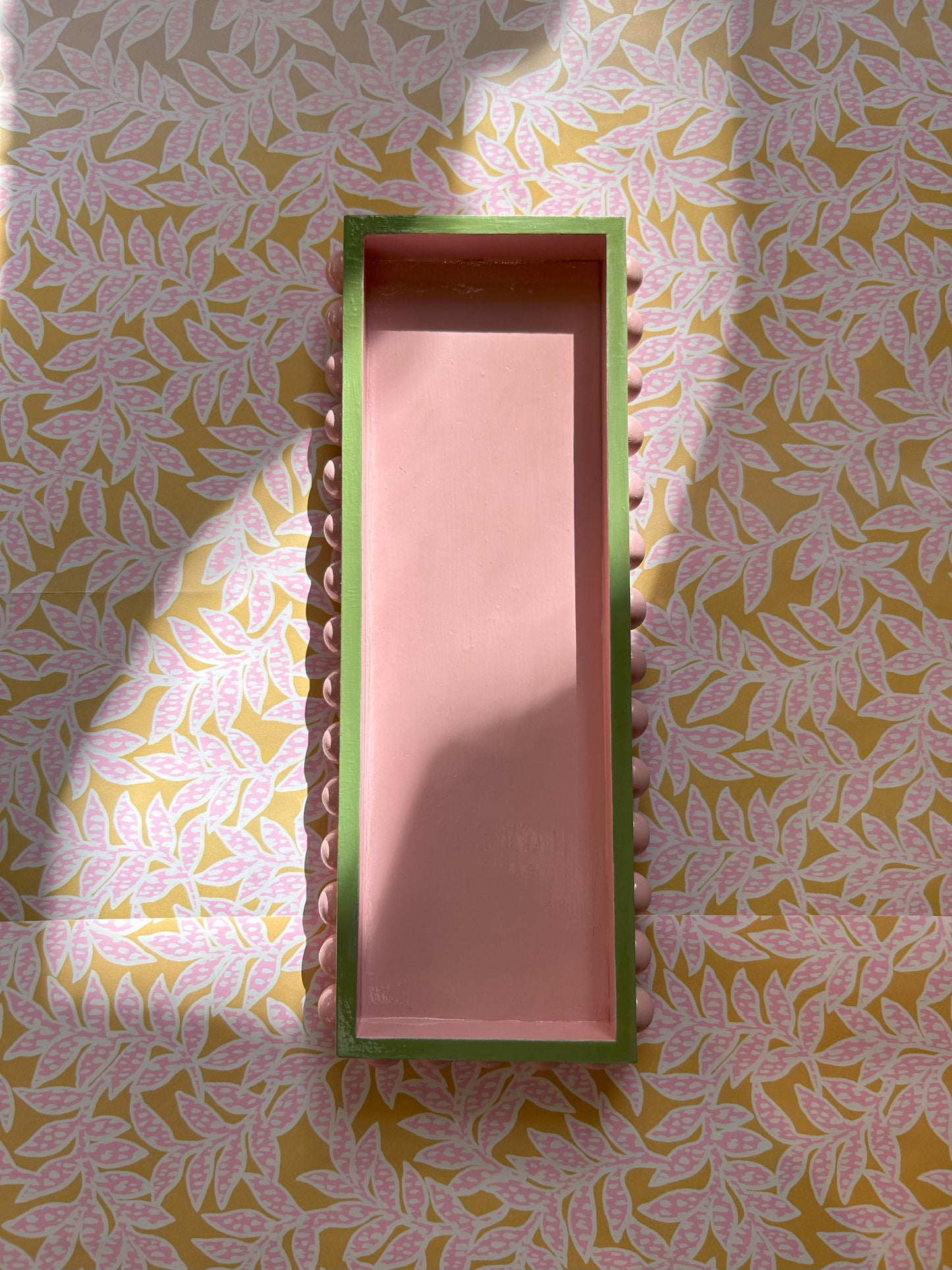 A rectangular decorative tray with a pink base and green edges, featuring wooden bobbins and a gloss finish, placed on a surface with a floral pattern.