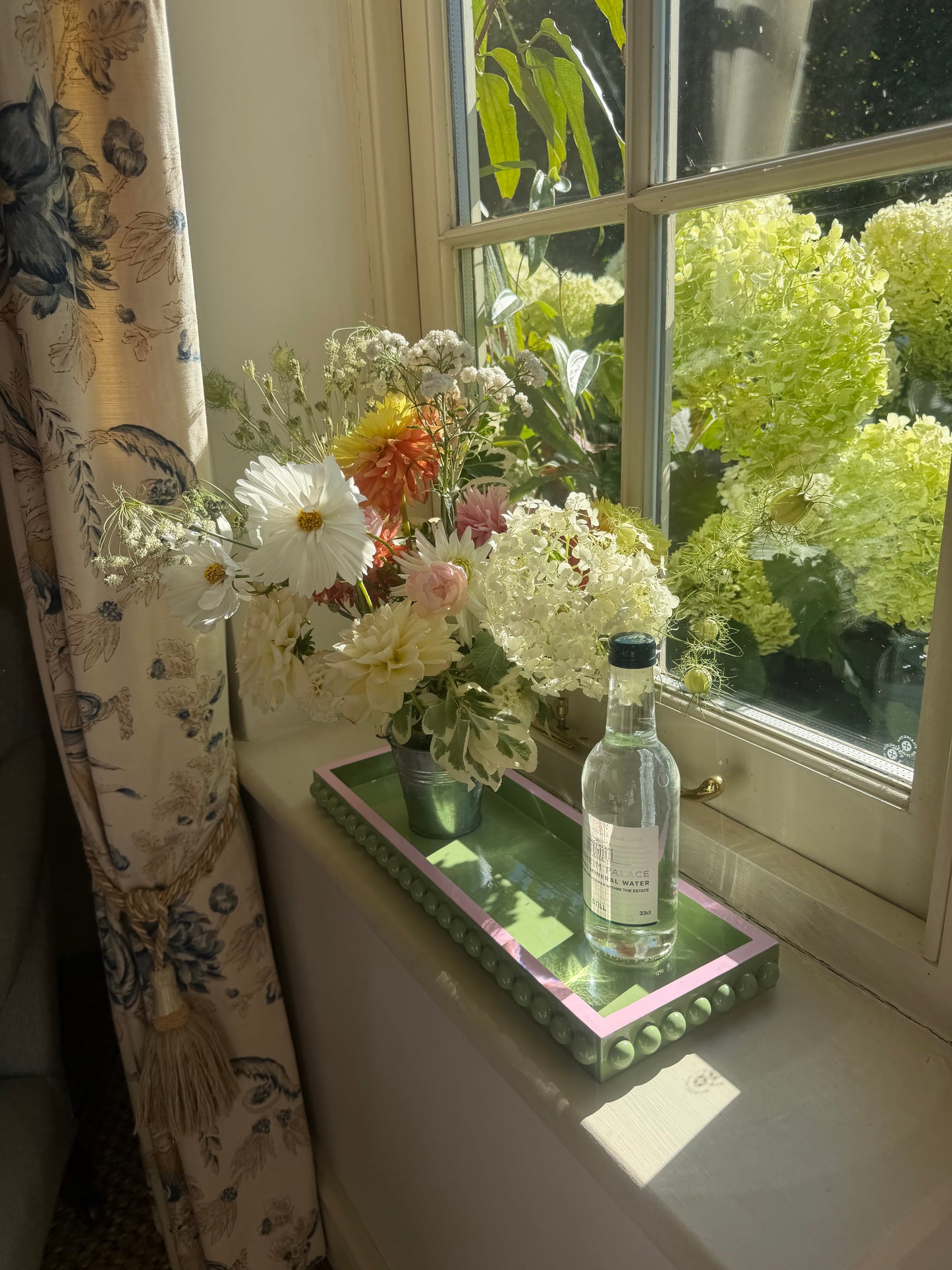 A clear glass bottle with a dark cap resting on a decorative pink and green tray, next to a bouquet of white and multicolored flowers on a windowsill with a window view of green hydrangeas in the background.