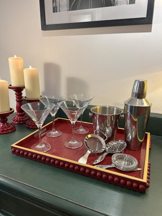 A rectangular, hand painted deep red coffee table tray with mustard yellow edge and handles, displayed with glassware and a shaker on top, set against a home backdrop.