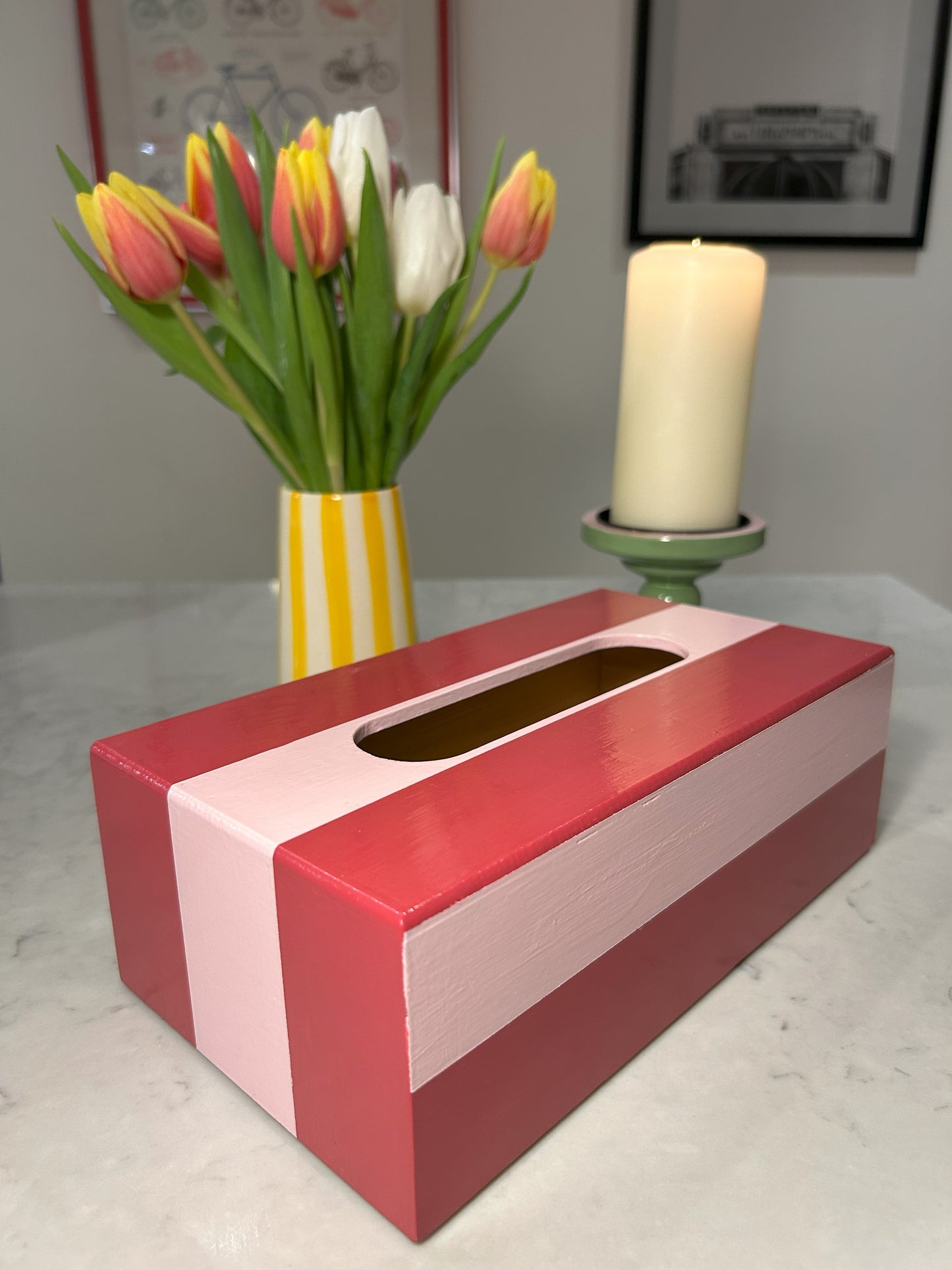 A hand painted red and pink striped tissue box on a table with a vase of tulips and a candle in the background.