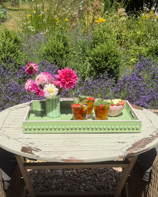 A distressed light green wooden outdoor table in a garden setting, styled with a matching green tray holding a vase of pink and white flowers and three small orange drinks, surrounded by wicker chairs.