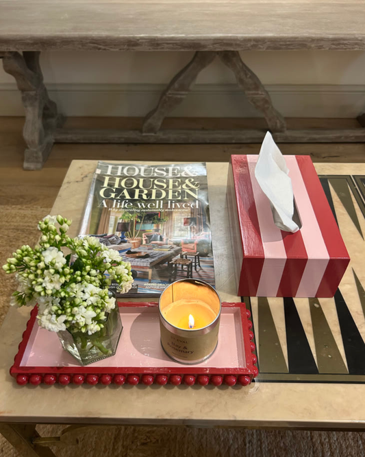 A styled tabletop scene with a red-and-white striped tissue box, a pink tray with a small flower arrangement and a lit candle, a magazine, and a black-and-gold geometric mat, set in a home interior.